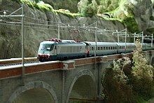 Maquette présentant une scène d'un train sur un viaduc, prise de trois-quarts avant. Le viaduc est gris, avec un parapet de briques rouges. Sur le viaduc, qui supporte une double voie, passe une locomotive électrique blanche et verte, tractant une rame de voitures de voyageurs de la même couleur. Le pantographe de la locomotive est levé, et touche la caténaire. L'arrière plan présente une structure rocheuse verticale, escarpée et grise, couronnée de végétation verte.