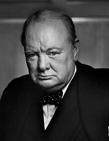 Churchill, 67, wearing a suit, standing and holding into the back of a chair