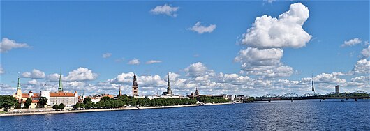 Blick über den vorderen Fluss zum Ufer mit Bäumen und vielen Häusern im Hintergrund. Einige Kirchtürme ragen hervor und eine Eisenbrücke führt rechts über den Fluss.