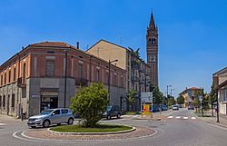 A small silver hatchback going around a roundabout in the foreground with a two-story tile-roofed building behind it. On the left side of a street leading into the roundabout from the background is brick campanile rising against a clear blue sky. There are signs in Italian and a crosswalk just behind the roundabout.