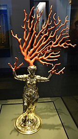 A gold figurine of a woman with raised arms. Her hands are small branches of real coral, and large branch of coral extends upwards from the top of her head.