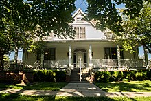 Photograph of a white two-story frame building with a front porch with green tree branches and dappled shade in the foreground