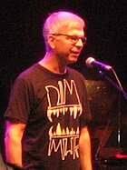 A gray-haired man with glasses and a black shirt standing in front of a microphone