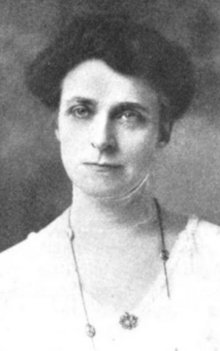 A white woman with dark hair in an updo, wearing a v-neck white garment and a necklace