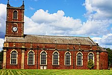 A brick church with stone dressings seen from the south. The west tower has a clock and pinnacles, and along the south face of the body of the church are Georgian-style windows.