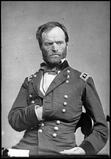 A black-and-white photograph of a man seated with his left hand inside of his jacket. His body faces the right diagonal and his head faces left.