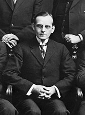 Black and white photo of a middle-aged seated in a chair, wearing a suit and tie