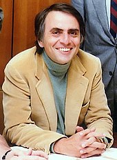 Un home de mitjana edat assegut davant d'una taula, vestit amb una jaqueta casual i un jumper. Ell està somrient i amb les mans plegades per posar-se per realitzar una fotografia; aquest és el físic Carl Sagan.