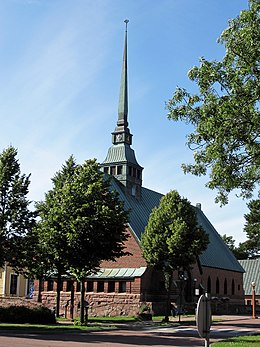 Bakstenen kerk met rondom een perkje met jonge bomen. De kerk heeft een grijs leistenen dak en opvallend is dat de zeer spitse toren niet aan het uiteinde van het dak staat maar op ongeveer een kwart vanaf de voorzijde.