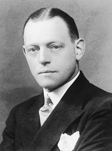 Sepia monochrome bust-length portrait of a clean shaven white male with short, slicked-back hair and a centre parting.