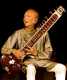 An old man sits on a platform and holds a long-necked lute while looking to the side.