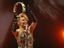 An image of Kylie Minogue, with one of her raising hands holding a tambourine. The microphone in a microphone stand is seen infront of Minogue.