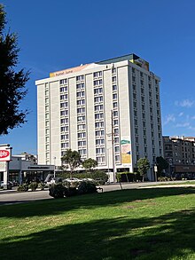 A hotel tower with a white exterior, against a blue sky, across a road; in the foreground, a grassy park lawn