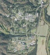 A top-down aerial view of a small, rural town. A river flanked by fields runs along the right side of the frame, while the rest of the town is surrounded by forest.