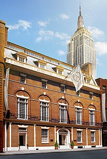 A multi-leveled brick building with many whitepaned windows with skycraper visible in the top right corner