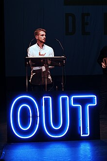 Adrián De La Vega parlant derrière pupitre. Devant lui, un grand signe "Out" en néon bleu