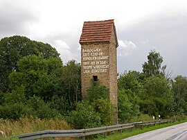 A small four-storey brick electrical tower with a red tile roof standing next to a road, with trees in the background. There is a wooden door to the right, and a window at first-floor level; the second storey and loft have no windows. The brickwork of the second floor bears a handwritten inscription, daubed with paint: "BARDOWIEK: SEIT 1292 URKUNDLICH ERWÄHNT 1977–'89 IM "DDR"-REGIME WIDERRECHTLICH ZERSTÖRT."