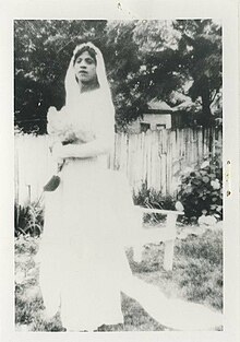 A black and white photo of the drag queen Sweet Evening Breeze in a wedding dress and veil holding a bouquet of flowers and standing in a garden.