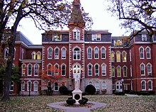 A statue sits in front of a large four-story red brick building. To the right of the entrance, the building goes in a little ways, and then back out. To the left of the entrance is a small two-story area connecting the main part of the building to the rest of it. Above and behind the two-story part is a four-story part of the building