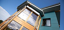 Der aus hellem Holz gefertigte Erker eines ansonsten blauen Hauses mit blauen Himmel im Hintergrund. Auf den Fensterscheiben spiegeln sich Cirrocumulus-Wolken.