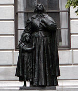 A photograph of a metal statue of a woman standing upright with her head tilted upward and her eyes looking up. She is dressed in a full dress, and beside her is a young girl who is clinging on to her.