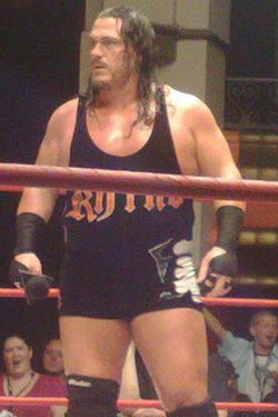 An adult white male wearing black standing in a wrestling ring.