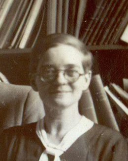 Photographie en noir et blanc d'une femme avec des lunettes et des cheveux courts devant une étagère avec des livres