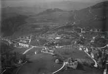 Schwarzweisses Luftbild mit Blick auf eine dünn besiedelte Hügellandschaft. An den hängen stehen vereinzelte Wohnhäuser. Es ist auch ein Bereich mit Gartenbeten erkennbar.