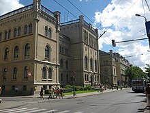 Seitliche Farbfotografie von einem grauen, dreigeschossigen Gebäude mit drei Vorbauten, die an den Ecken und in der Mitte sind. Leichte Ornamente sind an den Ecken und über den Rundbogenfenstern. Auf den Dächern sind Steingeländer, die von Eckpfeilern gestützt werden.