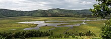 Ein mit Sumpfvegetation und Wasserflächen bedecktes Tal. Auf den Hügeln auf der anderen Seite stehen einzelne Häuser und an den Hängen kleine Landwirtschaftsflächen.