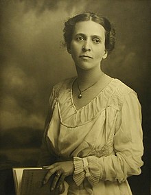 A sepia-toned photograph of a white woman, her dark hair parted center and dressed back; she is wearing a loose-fitting white dress with a v-neck and long sleeves, with a small pendant. She is holding a book in her hands.