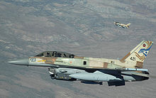 Two fighter jets with earth colored camouflage. The squadron emblem, a Star of David, and a design resembling a bird of prey are featured on the tail of one aircraft.