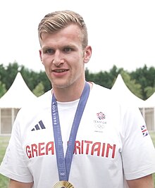 Tom Digby trägt ein weißes Hemd mit der Aufschrift Great Britain. Um den Hals trägt er ein blaues Band, an dem seine Goldmedaille befestigt ist, die aber nur zum Teil im Bild ist.