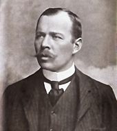 Head and shoulders portrait of a man with receding hair, heavy moustache, looking left from the image. He wears a high white collar, black necktie, dark waistcoat and jacket.