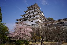 Château de pierre blanche de style oriental. Des cerisiers en fleurs sont également visibles.