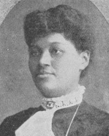 A Black woman a dress with a high lace trimmed collar with an attached white ruffle down the front