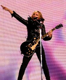 A female blond performer standing on a stage. She is wearing a black jacket and black pants with boots. Her hands are stretched upwards. She is looking to the right while holding an electric guitar in her left hand. Behind her, a violet screen is visible.