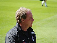 A middle-aged man, wearing a black tracksuit top. A grass field is visible in the background.