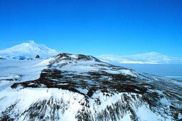 A rocky landscape covered in snow, with ocean in the background to the right and other snowy hills surrounded