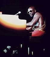 An adult male with sunglasses plays a piano under a spotlight on a darkened stage, 1973