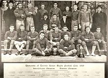 Twenty-five men, divided into three rows, pose. The majority are wearing football uniforms, except for four executives in suits. Two trophies are placed in front of the players.