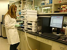 A woman in a lab coat takes glass vials out of a machine. A computer displays a spectroscopy graph.