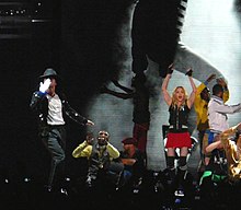 A blond woman dancing wearing a red shorts and black, sleeveless top. A man, wearing a black pant and jacket, with black hat on his head, dances beside her.