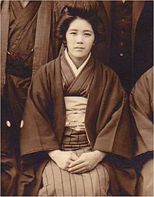 A black and white photo of a young woman, sitting, wearing a striped kimono and a jacket and a traditional hairstyle.