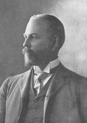 A black-and-white profile photograph of the head and shoulders of a middle-aged bearded man, facing left