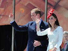 Catherine and William waving. She is wearing a red hat and a white dress along with a diamond brooch in the shape of a maple leaf.