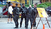 Four men wearing battle fatigues and holding guns and a crewmember wearing a white shirt, a hat and a walkie-talkie attached to its shorts stand on a set resembling a war torn city.