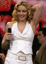 The front profile, up to the waist, of a middle-aged blond woman. She is wearing a white, sleeveless coat and white pants. Her hair is middle-parted and in locks around her face. She is holding a microphone in her right hand while her left hand is placed behind her head. She is smiling looking down. Behind her a video screen is visible whose picture is red.