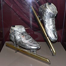 Metallic silver skates with golden blades in a glass case with the right skate being slightly elevated. There is a burgundy curtain behind the skates. The blades are much longer than the actual boot of the skates.