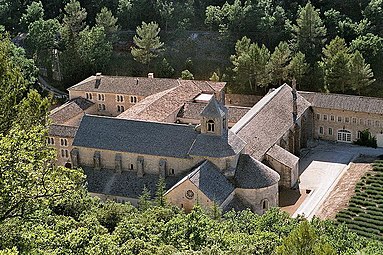 In a wooded valley is a large church with small windows and a square stone belfry. It is surrounded by ancient buildings arranged around courtyards, and a lavender garden.